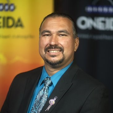 A middle-aged man in a blue shirt and tie and black suit coat is in front of an orange and purple banner with the word Oneida on it.
