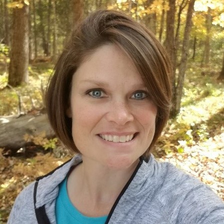 A woman with short, straight brown hair, and blue eyes is standing in a fall woods with yellow leaves. She's wearing a turquoise t-shirt and light grey quarter-zip top.