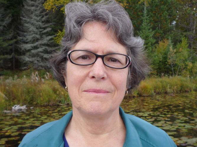 Headshot of a white woman with short dark hair and glasses with black frames wearing a teal shirt and standing in front of a wetland in fall