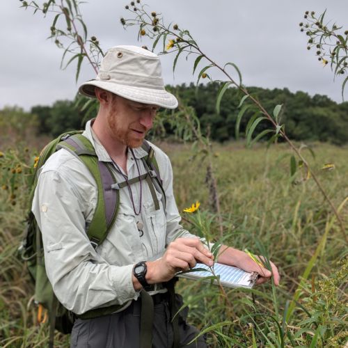 Man wearing a tan hat, tan shirt, and green backpack is standing in a wetland holding a clipboard and looking at plants.