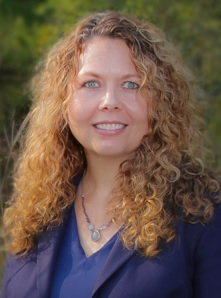 Smiling white woman with curly reddish-brown hair wearing a blue v-neck shirt and blazer standing in front of green vegetation