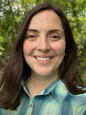 Smiling woman with shoulder-length dark brown hair and a plaid green and blue shirt standing in front of green vegetation