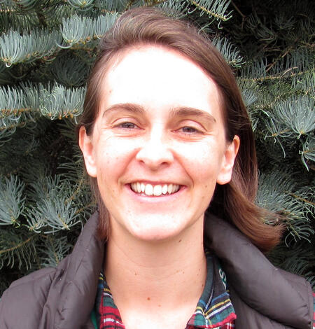 Smiling woman with shoulder-length dark brown hair and a plaid green and blue shirt standing in front of green vegetation