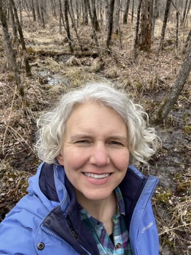 Smiling woman with silver hair and wearing a blue jacket is standing in a winter wetland.