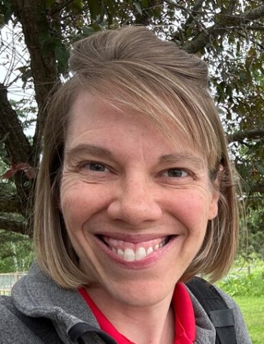 Smiling woman with light brown hair wearing a red shirt and grey fleece with trees and grass in the background
